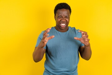 Young handsome man standing over yellow studio background Shouting frustrated with rage, hands trying to strangle, yelling mad.