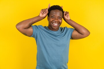 Funny Young handsome man standing over yellow studio background shows horns, fingers on head...
