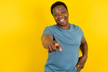 Young handsome man standing over yellow studio background pointing at camera with a satisfied,...