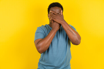 Young handsome man standing over yellow studio background Covering eyes and mouth with hands,...
