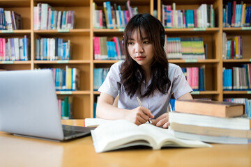 Young Asian female university student concentrate on studying, doing assuagement project in library with books and laptop on table