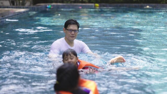 Asian family swimming in the pool on a happy vacation. A father teaches swimming to his child wearing a life jacket. Summer vacation activities