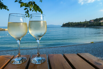 Two glasses of wine filled with white house wine, placed on a wooden table by the beach in Medveja, Croatia. The table is located in the shadow. Green leaves above. Clear and hot day. Summer holiday