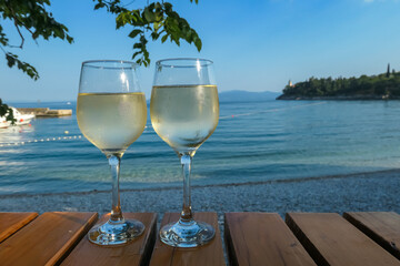 Two glasses of wine filled with white house wine, placed on a wooden table by the beach in Medveja, Croatia. The table is located in the shadow. Green leaves above. Clear and hot day. Summer holiday