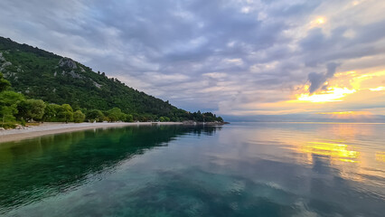 A stony beach along the shore of Medveja in Croatia. The Mediterranean Sea is calm and clear. There is a lush forest with a small town at the shore. The sky is painted yellow. Sunset time. Calmness