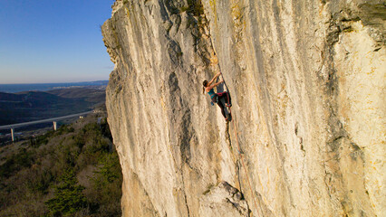 AERIAL: Mighty limestone cliff with young lady lead climbing a challenging route