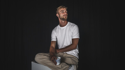 Young handsome blond man in casual t-shirt and beige pants on black background and sitting, posed seriously sipping a drink - obrazy, fototapety, plakaty