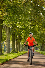 attractive senior woman cycling with her electric mountain bike in a beautiful old oak tree and chestnut avenue in Ludwigsburg, Baden-Wuerttemberg, Germany

