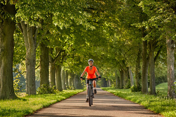 attractive senior woman cycling with her electric mountain bike in a beautiful old oak tree and...
