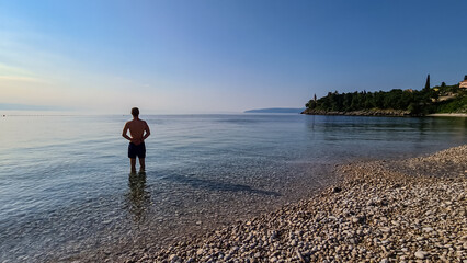 A man in swimsuit stands in the shallow water on a stony beach of Medveja in Croatia. The Mediterranean Sea is calm and clear. Some island visible. Clear, blue sky. Summer remedy. Holidays vibes.