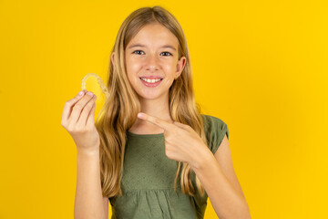 Beautiful caucasian kid girl wearing green dress holding an invisible aligner and pointing at it. Dental healthcare and confidence concept.