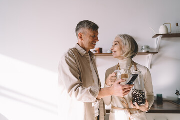Happy elderly couple with smartphone and jar in kitchen