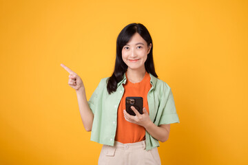 young 30s asian woman happy smile dressed in orange shirt showing smartphone with pointing finger hand gesture to free space isolated on yellow studio background. app smartphone concept