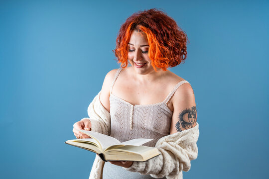 Modern tattooed non-binary redhead girl laughing with a book on blue background.