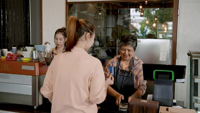 Elderly Asian mother standing chatting with customers ordering food drinks in store, customer asked about payment whether they could pay with credit card, mother handed cash machine customers tap pay.