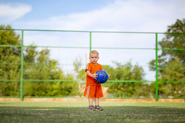 little blond boy in sports uniform posing with ball, child walking on playground on nature, active summer leisure concept