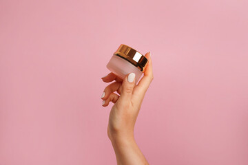 Female hand holding golden jar of cosmetic cream on pink background. Cosmetic beauty product branding mockup. Copy space
