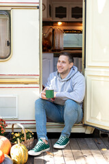 Campsite on caravan or camper van in forest. Rest man sitting on the doorstep of the motor home holding iron mug. Smiling handsome man sitting on the porch of his RV trailer holding a mug of coffee.	 - obrazy, fototapety, plakaty