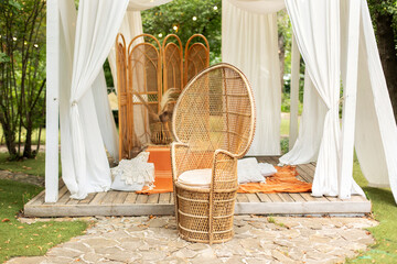 Rattan peacock armchair and decorative folding screen divider. Summer gazebo with flowing white curtains. Wedding boho decoration. Decor outdoor terrace with wicker furniture. Outdoor design of arbour © stock_studio