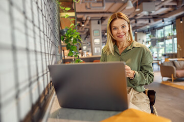 Smiling woman sales manager working on laptop in cozy coworking and looking at camera