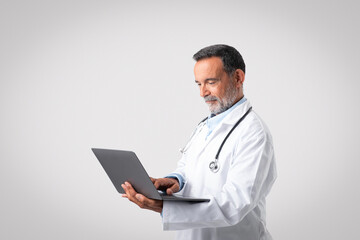 Happy concentrated caucasian senior therapist in white coat typing on laptop, doctor enjoy work