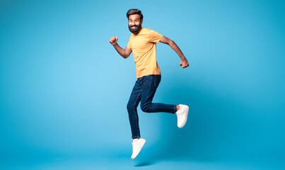 Fototapeta na wymiar Excited middle aged indian man running posing in mid air jumping over blue background, full length, free space