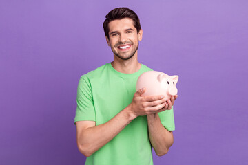 Portrait of satisfied cheerful guy with bristle wear green t-shirt holding pink small pig money box...
