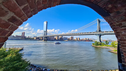 Obraz premium Iconic Manhattan Bridge connecting New York City's urban landscape with stunning skyline and architectural marvel, seen through a bricked wall. Tall skyscrapers. Famous spot for sightseeing
