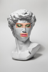 Replica of the head of an antique statue of David with a taped eyes and mouth with makeup on white background. Woman lips and eyes with makeup on statue.