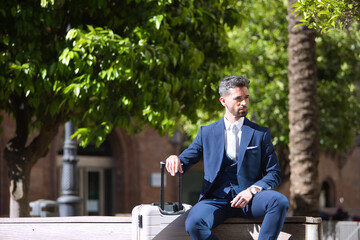 Obraz premium Young and attractive businessman, with beard and suit, sitting on a bench next to his suitcase waiting for a cab. Concept beauty, fashion, success, achiever, trend.