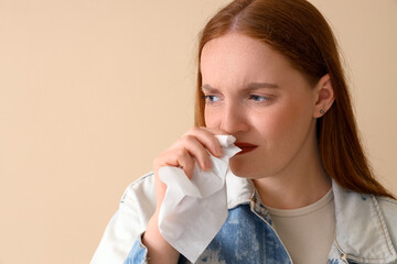 Allergic young woman sneezing into tissue on beige background, closeup