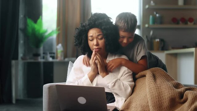 Exhausted young curly woman mother get struggles while working on laptop computer and her little son boy child appears to cheer her up and hug her tightly at home Family support concept