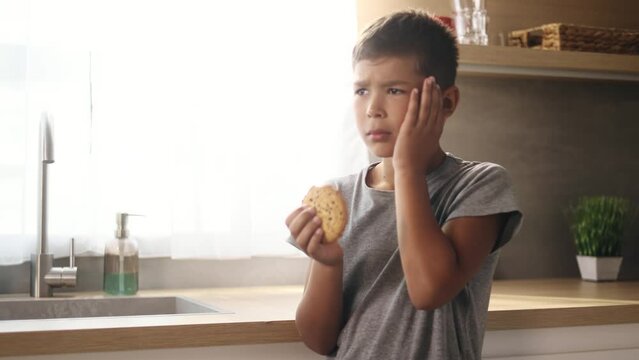 Portrait of cute little boy child eating cookie and having toothache standing at kitchen at home Sad kid feeling pain in tooth because caries gum disease or sensitive teeth indoors