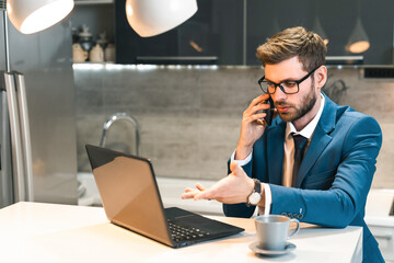Dedicated young businessman wearing suit and glasses holding smartphone, having a call in home office, working, consulting client, providing business service.
