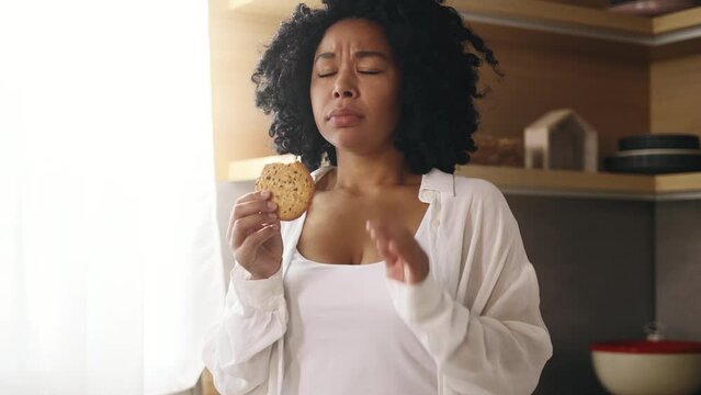 Portrait of young curly woman eating cookie and having toothache standing at kitchen at home Sad female feeling pain in tooth because caries gum disease or sensitive teeth indoors