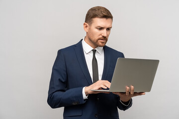 Serious frowning businessman in suit looking at laptop screen on grey background. Office work, job. Problems, troubles at work, mistakes, errors in documents, bad news, negative reactions concept.