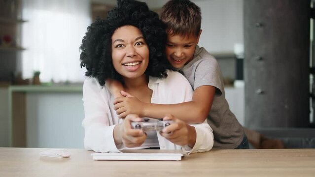 Camera view of excited beautiful curly woman gamer sitting at desktop and playing in video games on digital console while her son supports and encourages mother at home together Happy family concept
