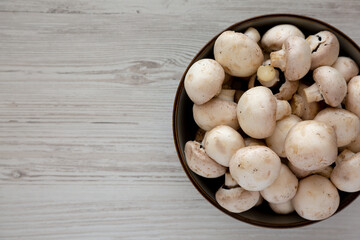 Raw White Champignon Mushrooms in a Bowl on a white wooden background, top view.