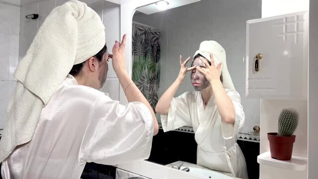 Young woman wearing towel on head and bathrobe putting clay mask on face.