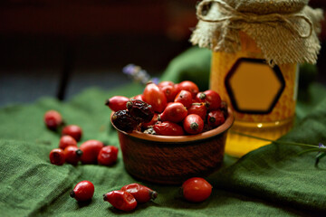 Rosehip berries in a bowl and scattered on a green tablecloth. A jar of light honey is in the background. Hot beverage recipe. Lifestyle photo. Autumn mood. Concept of a healthy diet.
