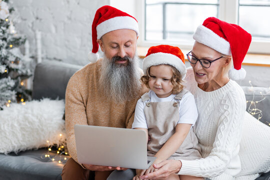 Concept of spending vacations or holidays at grandparents. Attractive active grandfather and grandmother, senior man and woman, with grandchild doing video call with parents on laptop or smartphone