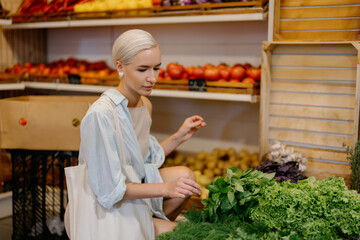 Woman is browsing and selecting various greens at a grocery store. She is wearing a light blue shirt and holding a white tote bag. Variety of healthy food options available at the store