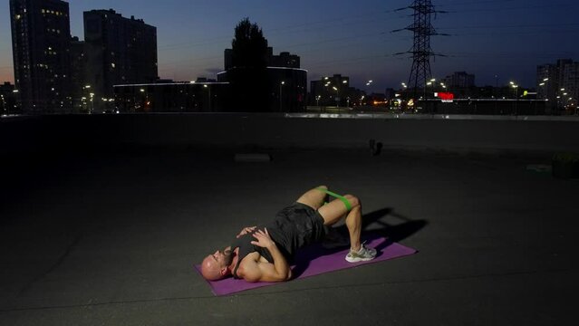 Fitness classes on a mat on the roof of the house at night. A handsome man performs yoga exercises in a city urban landscape. High quality 4k footage