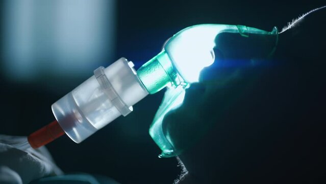 A female patient in medical mask is breathing oxygen support in a hospital next to heart rate machine. Unhealthy woman in coma. Getting mechanical ventilation. Concept of pandemic and therapy.