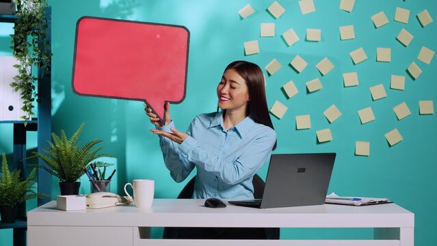 Happy cheerful asian businesswoman holding red speech bubble sign of empty space for text and showing ok hand gesture. Smiling employee presenting thought bubble cardboard in colourful workplace