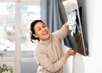 Hardworking asian woman wipes the dust from the TV with a rag, tidying up the apartment