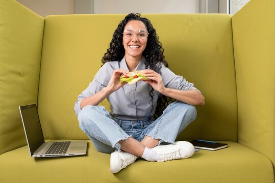 Lovely young Hispanic woman sitting in a coworking space with a laptop holding a sandwich in her hands and ready to eat. Cute curly lady took a break from work or study and starts enjoying food