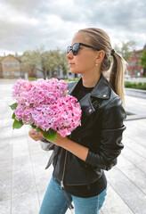 Fototapeta premium Beautiful caucasian girl with blond hair in a black jacket and blue jeans with a bouquet of pink hydrangeas and with the sunglasses with a city view in the background.