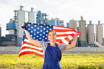 Happy young woman worker holding big flag of the USA against background of factory