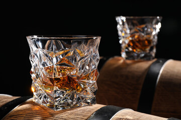 Glasses of tasty whiskey on wooden barrels against black background, closeup